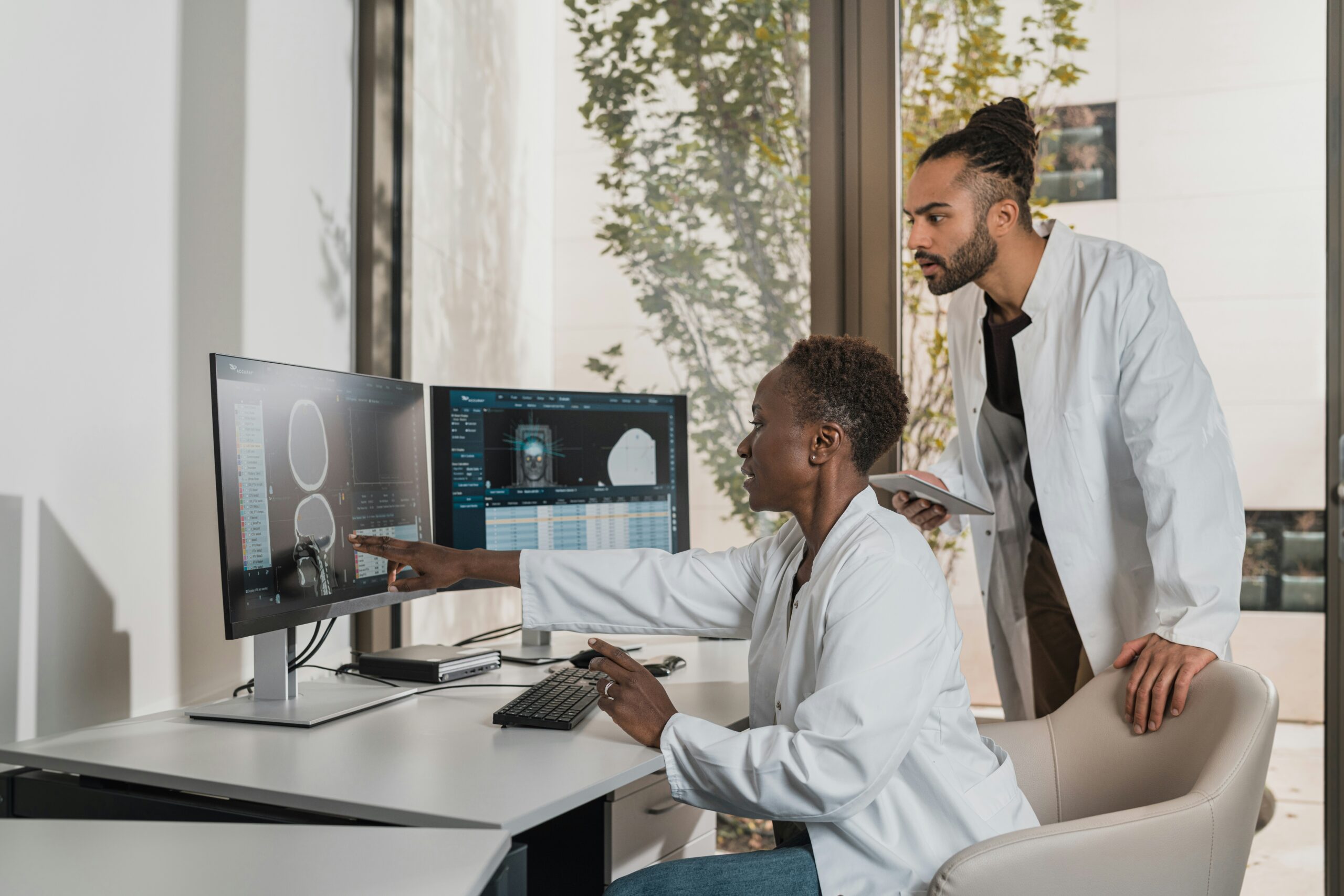 Two medical professionals in white coats review brain MRI scans on computer monitors in a bright office setting.