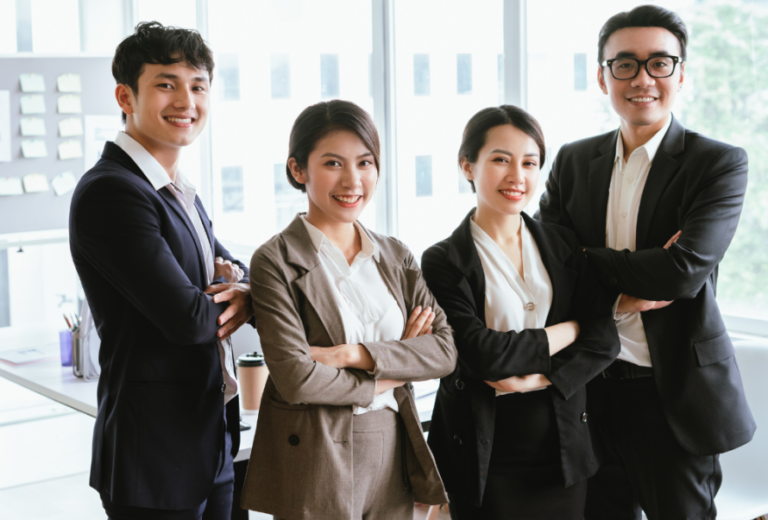 Four professionals in business attire pose with arms crossed in a bright office, smiling at the camera.